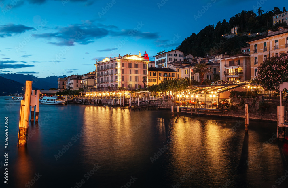 Obraz premium Bellagio's waterfront in Lombardy, Italy, glows with warm evening lights. Charming buildings line the shore, creating a picturesque scene as the water reflects the vibrant ambiance