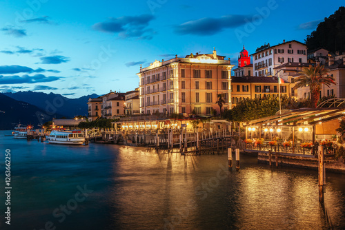 Scenic view of Bellagio at dusk with illuminated buildings along Lungolario Guglielmo Marconi. Calm waters of Lake Como reflect the vibrant evening lights, creating a serene atmosphere
