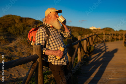 Elderly Bearded Man in His 70s Enjoying a Hot Drink from a Mug While Leaning on a Wooden Fence at Sunset photo