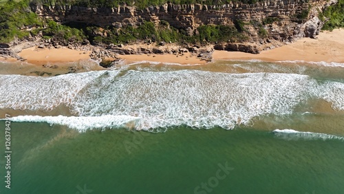 Drone shot aerial view of waves crashing onto the shore near the base of a cliff in summer in Australia 
