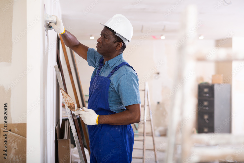 Painter in blue overalls leveling plaster on the wall with a spatula Painter in blue overalls leveling plaster on the wall with a spatula