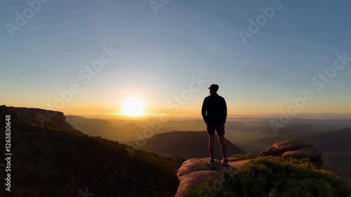 A solo traveler standing on the edge of a cliff, looking out over a breathtaking valley at sunrise.