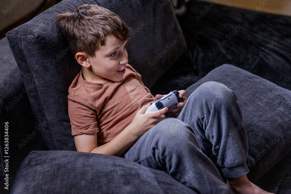 Fototapeta premium Boy is sitting on sofa at home and playing video games with his joypad.