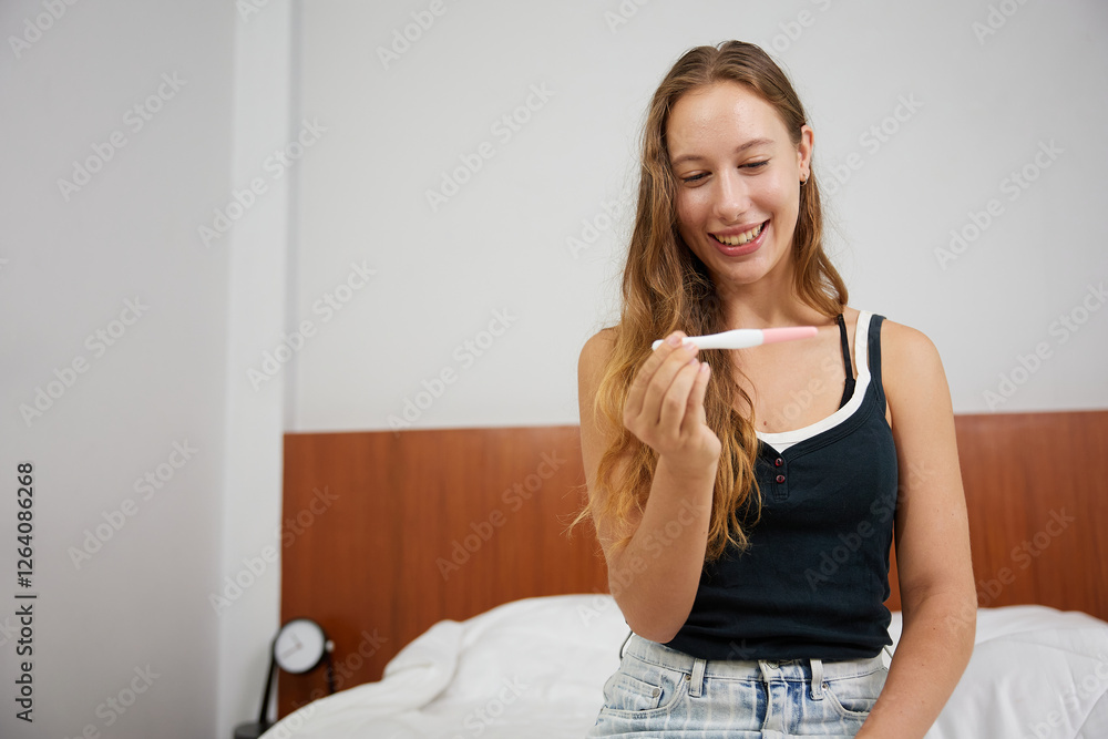 young woman looking at pregnancy test and smiling while sitting on bed at home