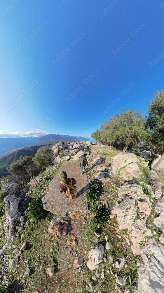 Vertical video. Wide-Angle View of Two Hikers Walking Along a Rocky Trail on a Mountain Ridge Surrounded by Trees and Overlooking a Valley Vertical video. Wide-Angle View of Two Hikers Walking Along a Rocky Trail on a Mountain Ridge Surrounded by Trees and Overlooking a Valley