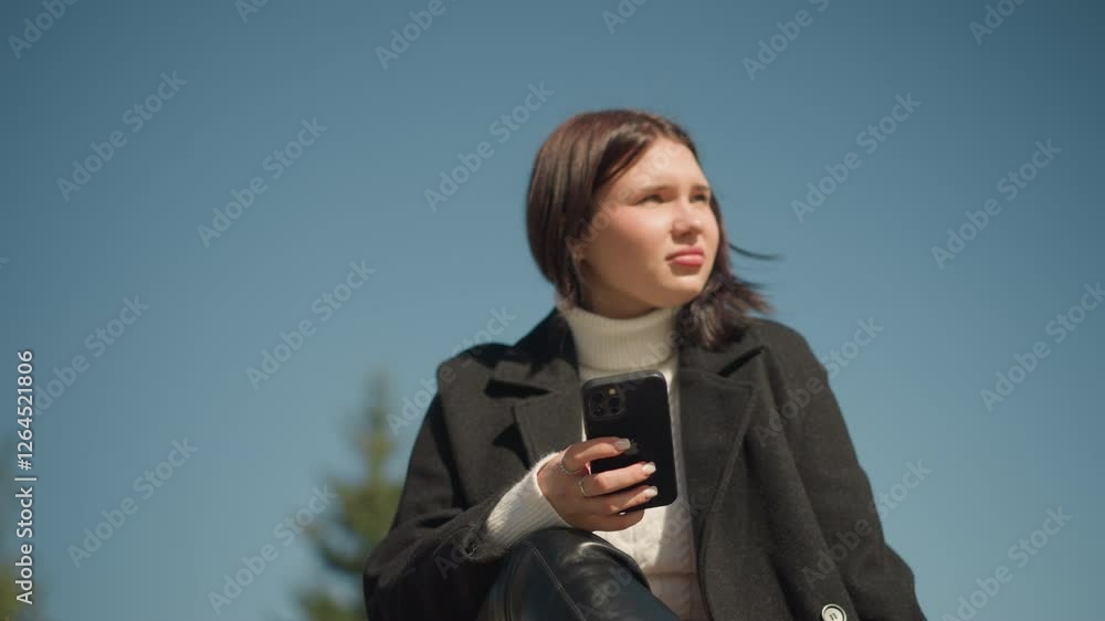 Close-up of young woman in black coat and white sweater sitting on bench outdoors, looking into distance with smartphone in hand, bright sunlight illuminating her face and green background Close-up of young woman in black coat and white sweater sitting on bench outdoors, looking into distance with smartphone in hand, bright sunlight illuminating her face and green background