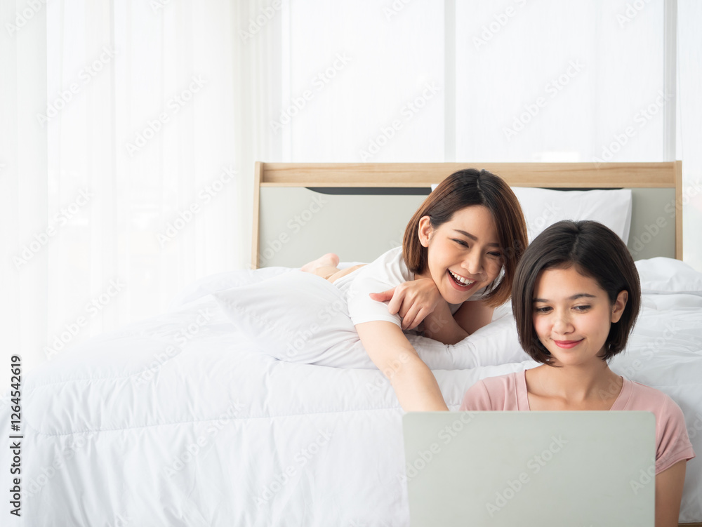 Asian two young women friends relax in bed, smiling and enjoying time together while using a laptop, sharing a moment of online entertainment Asian two young women friends relax in bed, smiling and enjoying time together while using a laptop, sharing a moment of online entertainment