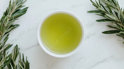 A bowl of green oil surrounded by fresh rosemary on a marble surface. photo