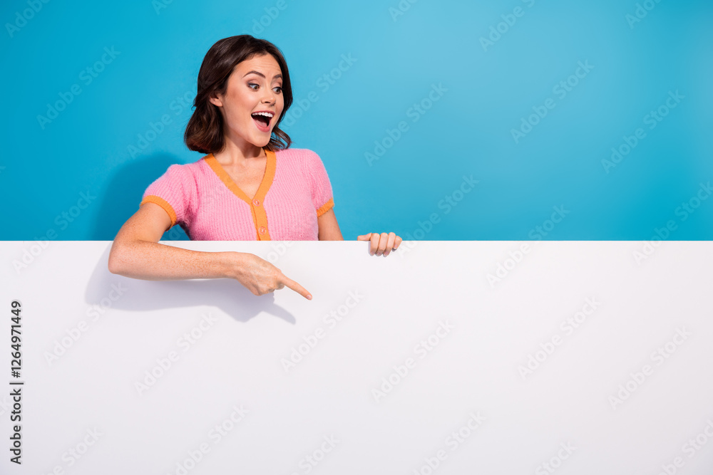 Excited young woman in pink top points downwards at empty white space against a vibrant blue background Excited young woman in pink top points downwards at empty white space against a vibrant blue background