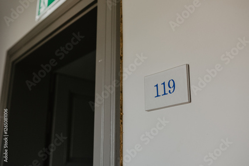 Open door leading to a dimly lit room with a white wall featuring a blue-numbered room sign 