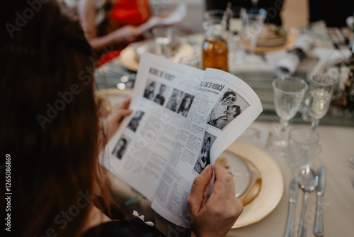 A guest at an elegantly set dining table attentively reads a black-and-white printed wedding newspaper featuring vintage photographs and articles, surrounded by fine glassware, gold-rimmed plates