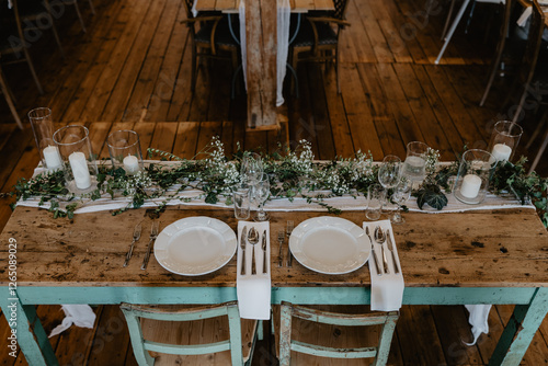 A beautifully arranged rustic wedding reception table with vintage wooden chairs, elegant white plates, polished silverware, and delicate floral greenery intertwined with candles in glass holders