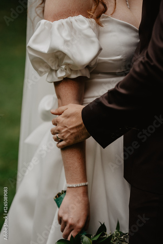  A groom in a dark suit tenderly holding the bride’s arm, showcasing their wedding rings, as she wears a white satin dress with elegant puff sleeves, a delicate diamond bracelet, and holds a bouquet 