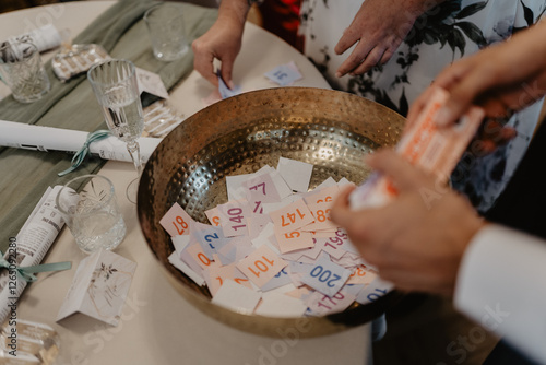 A close-up of a golden raffle bowl filled with colorful numbered tickets at an elegant event, where guests' hands are seen drawing and organizing tickets on a beautifully decorated table 