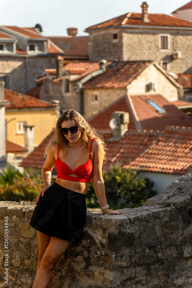 A stylish woman poses against the historic stone walls of a European old town. The warm sunlight highlights the rustic rooftops, creating a travel and fashion-inspired scene. A stylish woman poses against the historic stone walls of a European old town. The warm sunlight highlights the rustic rooftops, creating a travel and fashion-inspired scene.