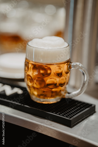 A traditional dimpled glass mug filled with golden amber beer with a thick frothy head placed on a black bar mat in a warmly lit pub setting with blurred bokeh in the background