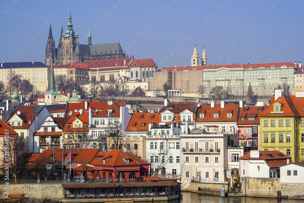 Obraz premium The view shows colorful buildings with red rooftops, with Prague Castle towering in the background under a clear blue sky