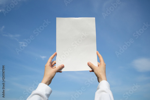 Hands holding up a blank sheet of paper outdoors, on a blue sky background.