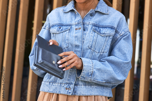 Woman opening stylish leather wallet, wearing casual denim jacket, elegant outdoor fashion, accessory and style representation concept