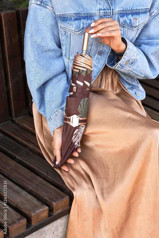 Woman holding stylish closed umbrella sitting on wooden bench wearing denim jacket and beige dress outdoor autumn fashion concept