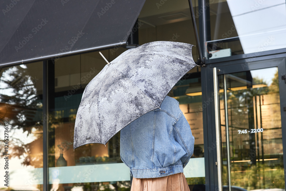 Stylish person with snake pattern umbrella, wearing denim jacket, outdoor cafe setting, standing near glass door, rainy weather concept