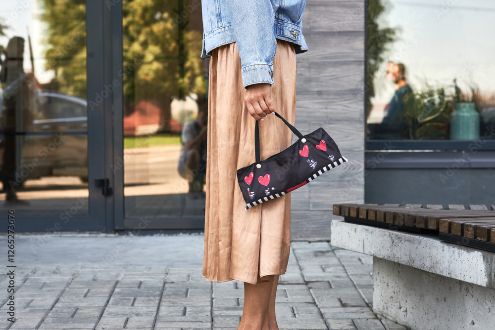Woman stylishly holds decorative purse, colorful hearts, wearing denim jacket and skirt, urban street concept