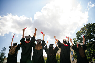 Rear portrait of group students raise fists hold diploma mortarboard gown graduate college building outdoors