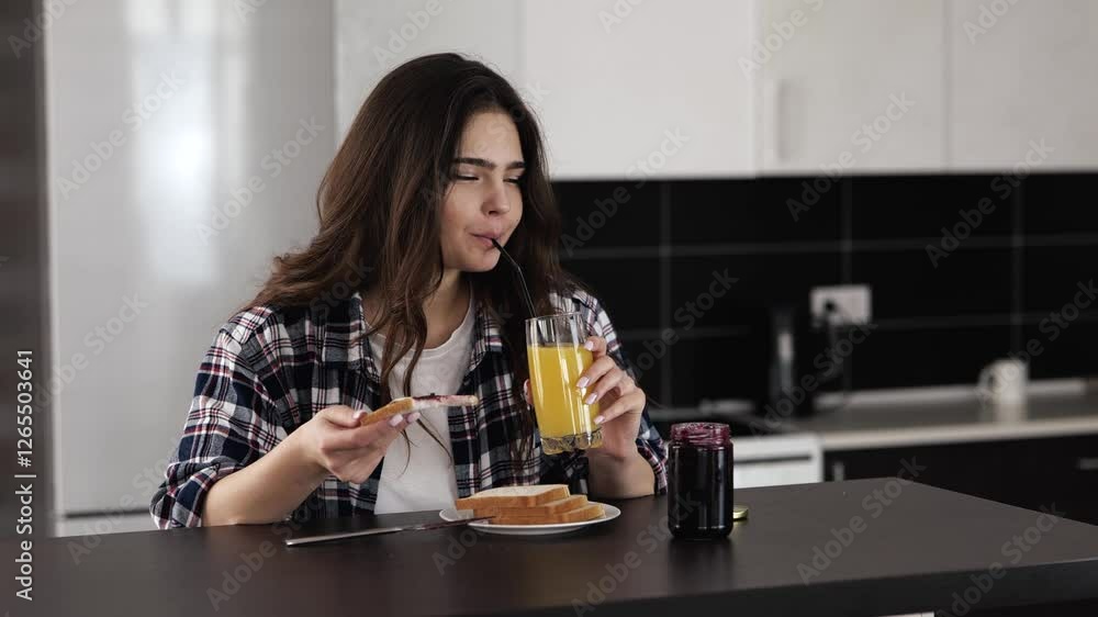 Young woman in kitchen during quarantine. Girl eating bread with jam and smile. Cheerful female person drinking orange juice. Enjoying breakfast in morning. Young woman in kitchen during quarantine. Girl eating bread with jam and smile. Cheerful female person drinking orange juice. Enjoying breakfast in morning.