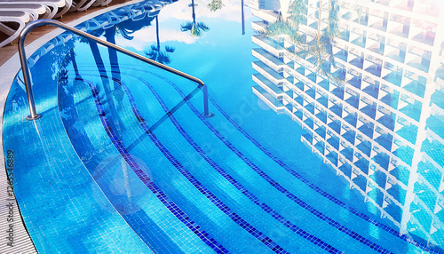 Blue fresh swimming pool with  handrails and stairs  into the water, luxury hotel and trees reflected in the water in sunny summer day. Summer, vacatio concept. photo