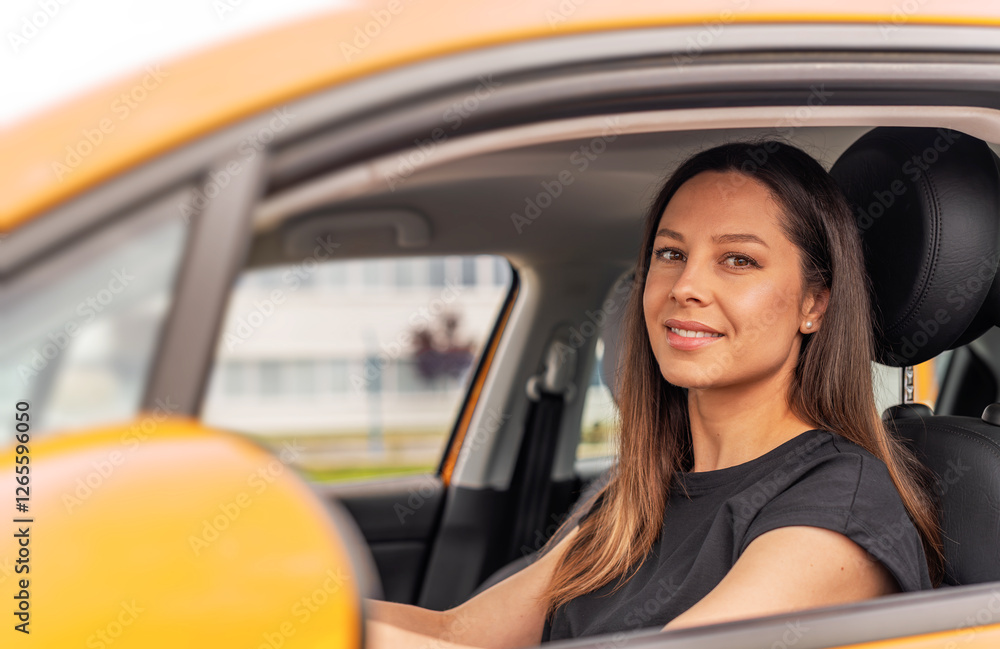 A young beautiful woman with long brown hair and a bright smile sits confidently in the drivers seat of a yellow car, holding the steering wheel and looking outside with a happy expression.
