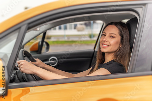 Obraz na płótnie A young beautiful woman with long brown hair and a bright smile sits confidently in the drivers seat of a yellow car, holding the steering wheel and looking outside with a happy expression. w salonie