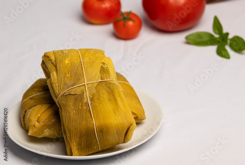 humita en hoja cerrada sobre un plato blanco, sobre un mantel blanco, al fondo tomates maduros de diferentes tamaños y una rama de albahaca photo