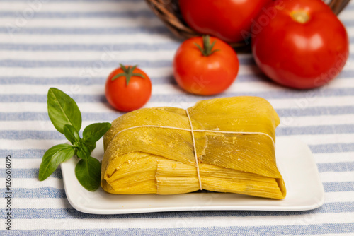 humita envueltas en hojas de choclo cerrada con pitilla sobre un plato blanco decorado con una hoja de albahaca, sobre un mantel con lineas, al fondo tomates maduros que salen de un canasto photo