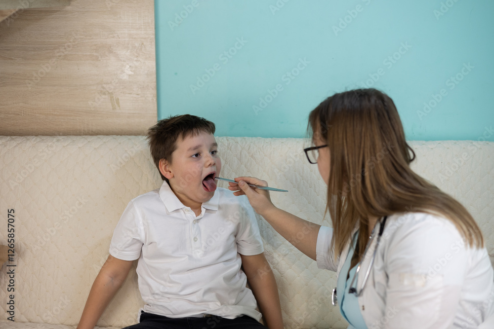 A female doctor uses a tongue depressor while examining the throat of a sick boy sitting on a couch at home during a house call. The doctor provides help and support to the child. A female doctor uses a tongue depressor while examining the throat of a sick boy sitting on a couch at home during a house call. The doctor provides help and support to the child.