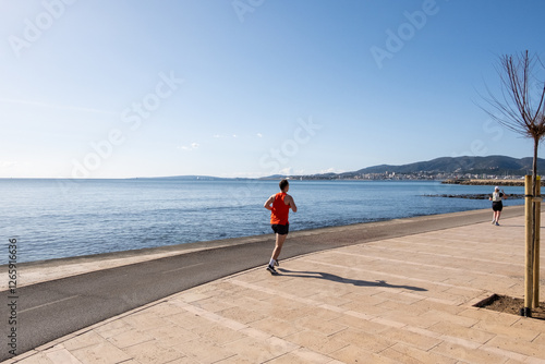 man from behind in a red shirt doing running exercise on the beach promenade during the beautiful sunny day