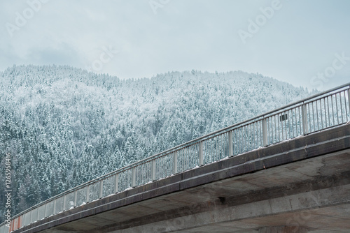 Obraz na płótnie Concrete Bridge with metal railing is visible crossing the river and snow capped hills with forest seen in the background w salonie