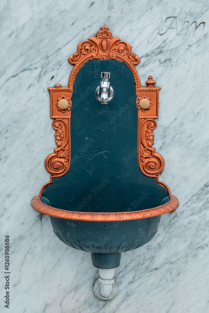 Front view of a vintage ornamental water basin and faucet mounted on a marble wall. Golden ornaments at side.