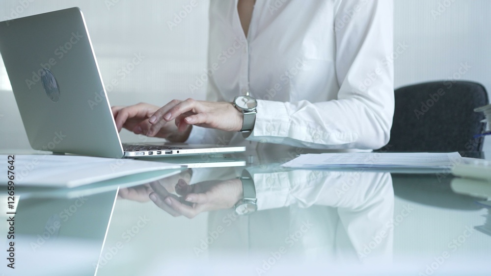 Businesswoman in white blouse typing on laptop at glass desk while sitting in bright office, showcasing productivity and modern work environment. Businesspeople concept