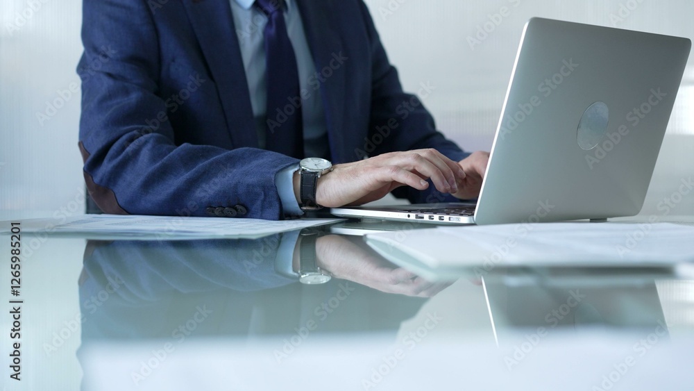 Businessman in blue suit is typing on laptop at glass desk while sitting in bright office, showcasing productivity and modern work environment. Businesspeople concept Businessman in blue suit is typing on laptop at glass desk while sitting in bright office, showcasing productivity and modern work environment. Businesspeople concept