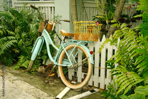 Old aqua blue beach cruiser bike with whitewall tires, bell and a basket in the front leaning up against white picket fence beside a house porch. Sitting on a wood panel and concrete driveway. No peop