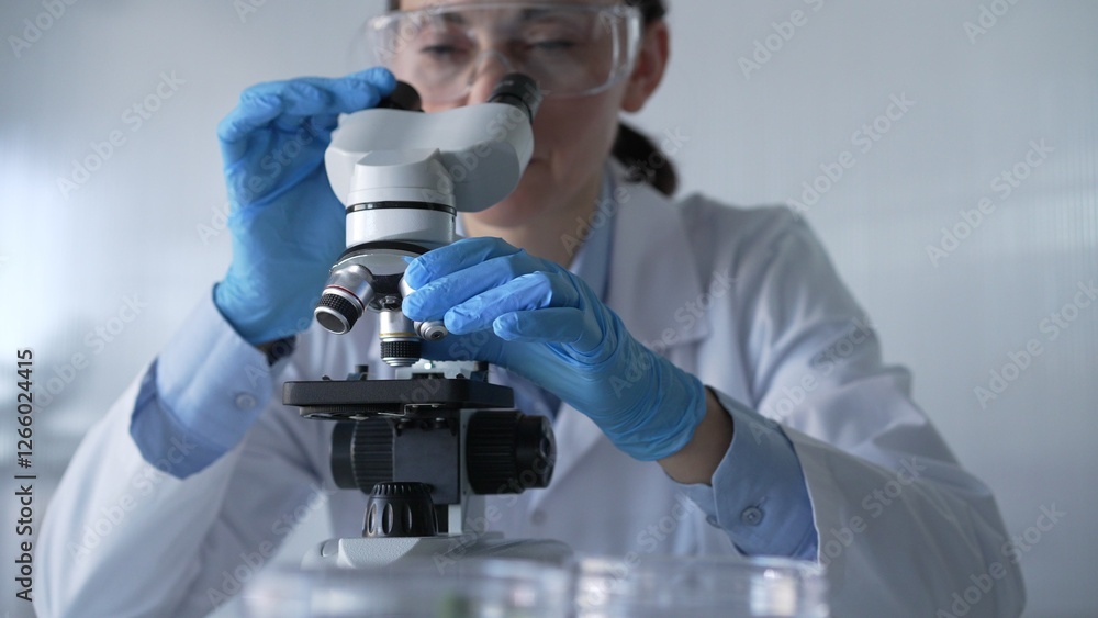 Woman scientist wearing lab coat, white protective gloves and glasses, is using microscope in modern laboratory setup, portrait view. Low key lighting photo. Science and Medicine Woman scientist wearing lab coat, white protective gloves and glasses, is using microscope in modern laboratory setup, portrait view. Low key lighting photo. Science and Medicine