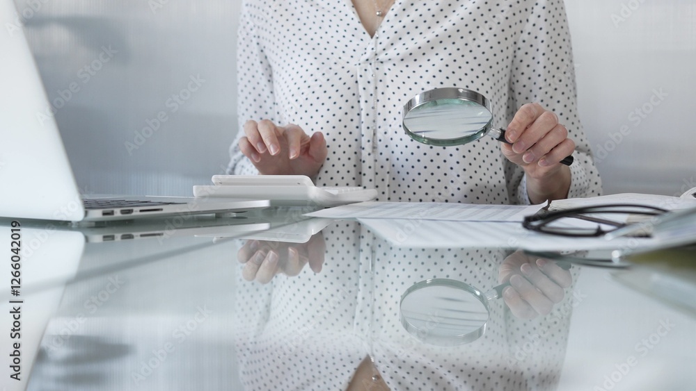 Businesswoman carefully reviewing financial reports with magnifying glass and calculator, conducting audit and analyzing data. Taxes, audit in business