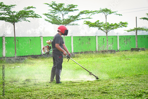 A man in a helmet mows a grassy field near a green wall. He is using a gas-powered trimmer, and the image shows him hard at work.
