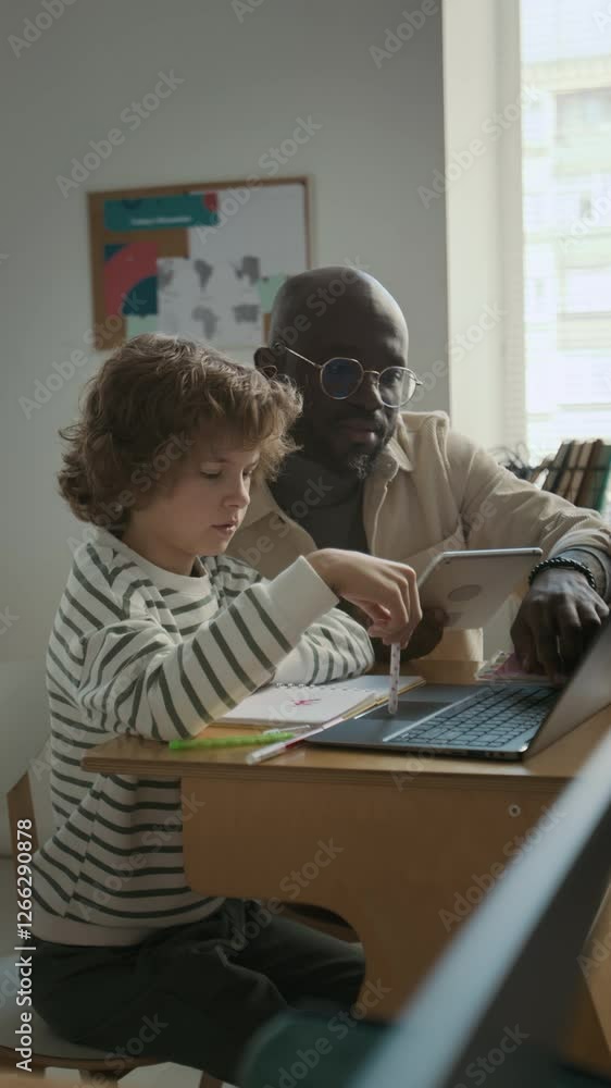 Vertical shot of African American male teacher sitting at desk together with student, helping with task during class in modern school Vertical shot of African American male teacher sitting at desk together with student, helping with task during class in modern school