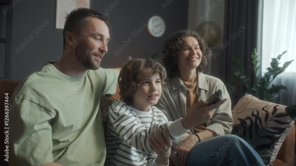 Joyful parents and little son sitting together on couch in cozy living room, watching TV and laughing, boy holding remote control and switching channels Joyful parents and little son sitting together on couch in cozy living room, watching TV and laughing, boy holding remote control and switching channels