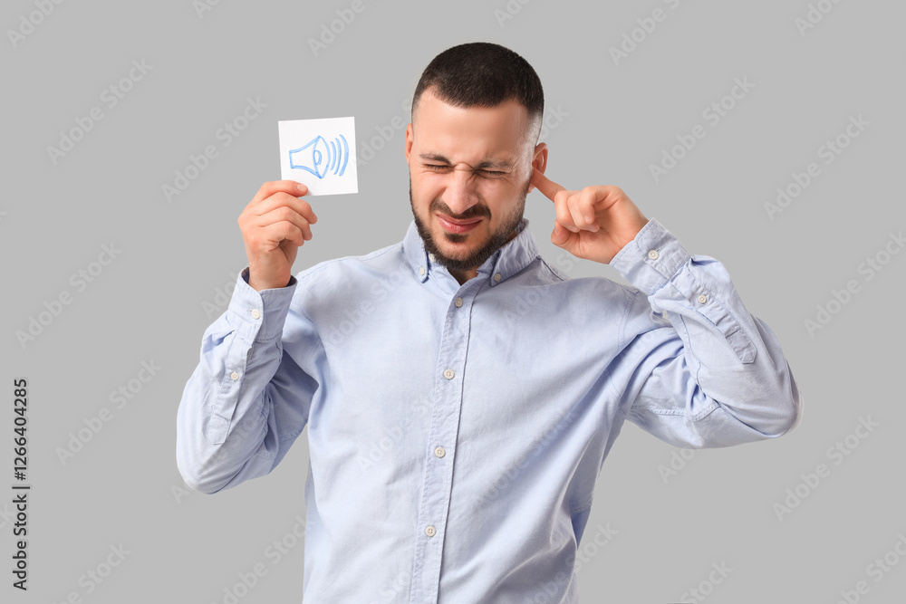 Young businessman with paper speaker icon suffering from loud noise on grey background Young businessman with paper speaker icon suffering from loud noise on grey background