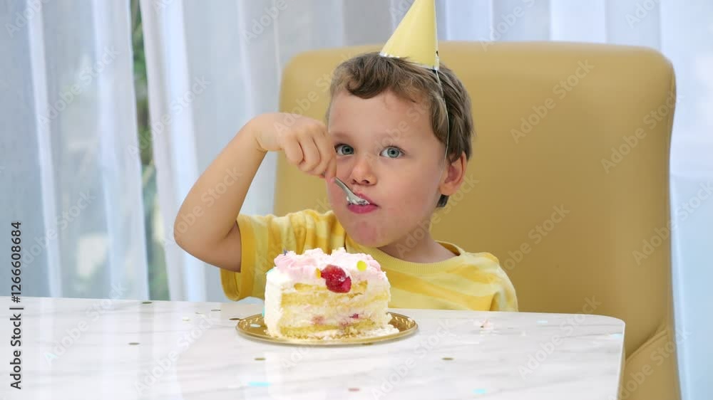 Cheerful toddler wearing colorful party hat enjoying sweet birthday cake slice. Sitting by the table, smiling kid take piece of sweet food by spoon and enjoy its taste.