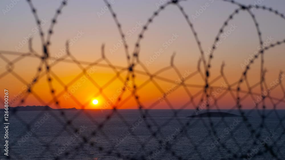 Orange sunset behind barbed wire over calm sea. Barbed wire fence dominates the foreground, obscuring a tranquil ocean sunset view, evoking longing, and the contrast between freedom and captivity Orange sunset behind barbed wire over calm sea. Barbed wire fence dominates the foreground, obscuring a tranquil ocean sunset view, evoking longing, and the contrast between freedom and captivity