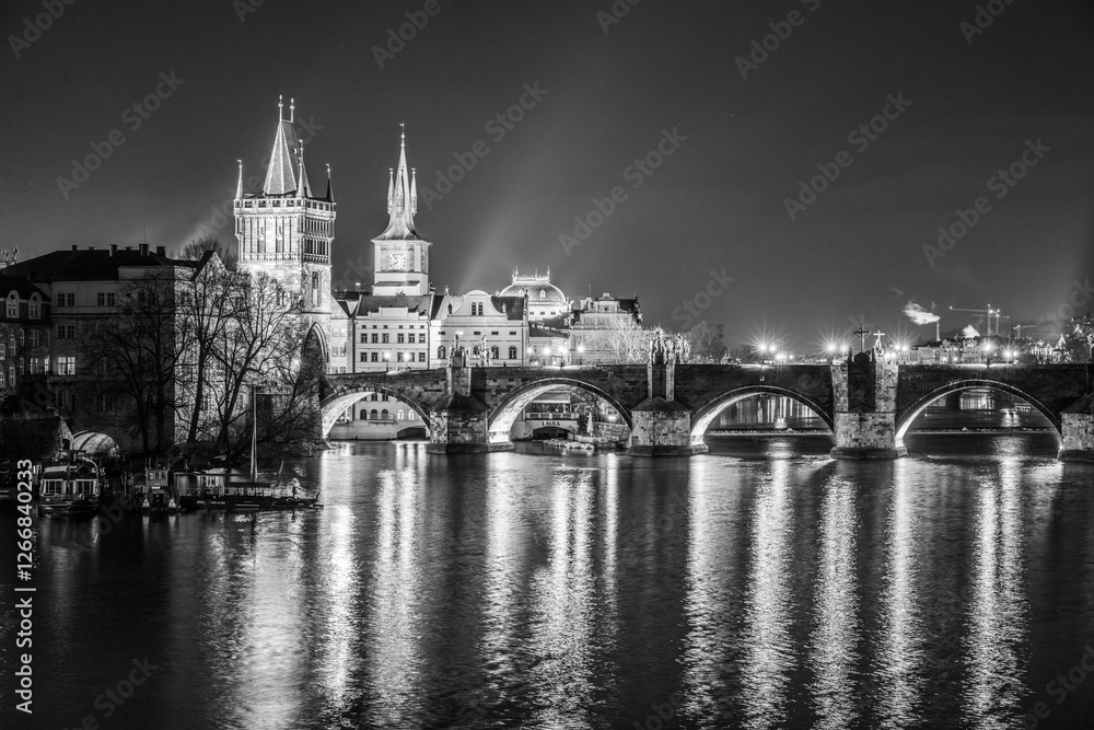 Fototapeta premium Illuminated towers and arches of Charles Bridge reflect in the tranquil Vltava River during a serene night in Prague. The city lights create a stunning nighttime ambiance.