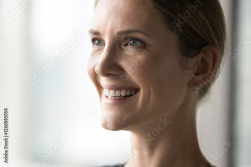 Close up face attractive young 35s woman with positive expression looks into distance, posing indoor. Profile of beautiful authentic optimistic female, having wide charming candid smile staring aside photo